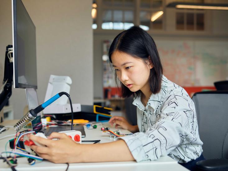 Side profile of Tiffany Louie sitting and leaning onto a table with a monitor, soldering gun, colorful wires and folders splayed next to her