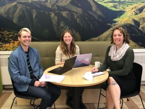 Emily Levenson sitting at a round table with two coworkers in New Zealand with their laptops open with a mountainous region scenery on the wall