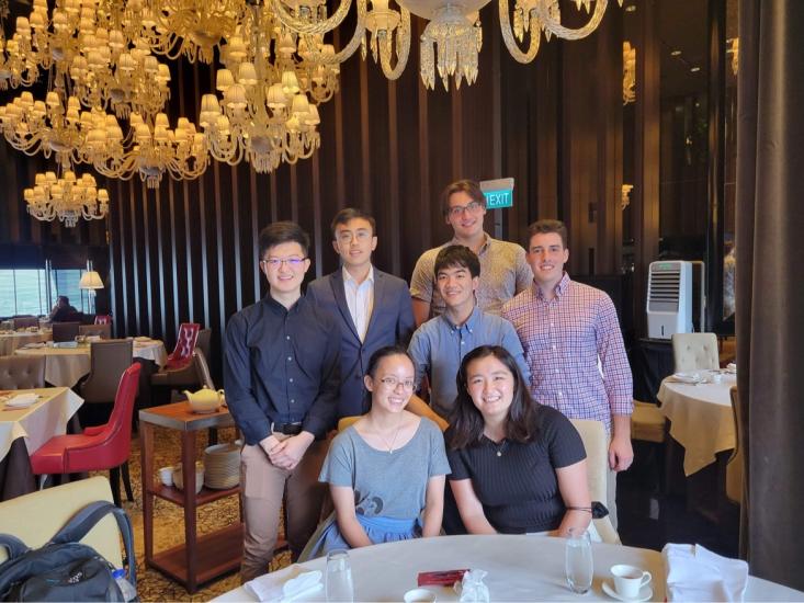 Joseph Cahaly and 6 other MIT students pictured in a restaurant in Singapore with chandeliers and white-covered table cloths a cart at the back with a white teapot on it
