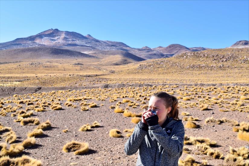 Kira Buttrey drinking from a cup in Chile's Atacama, a desert brown plants across a plants and a mountainous region in the background