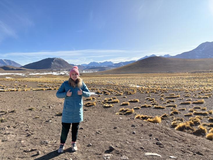 Mikayla Britsch dressed in winter wear in Atacama Desert in Chile with mountainous range in the background and brown plants around
