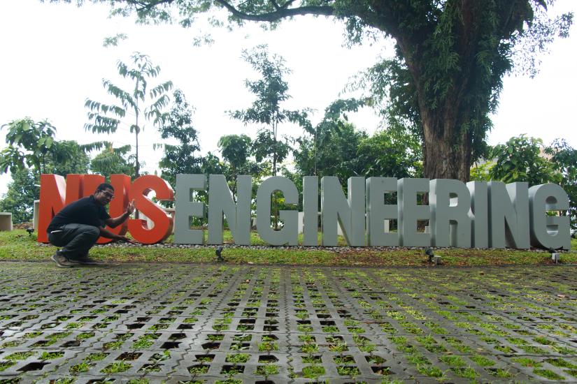Zachary Francis crouching down in front of the NUS Engineering sign in Singapore with trees in the background