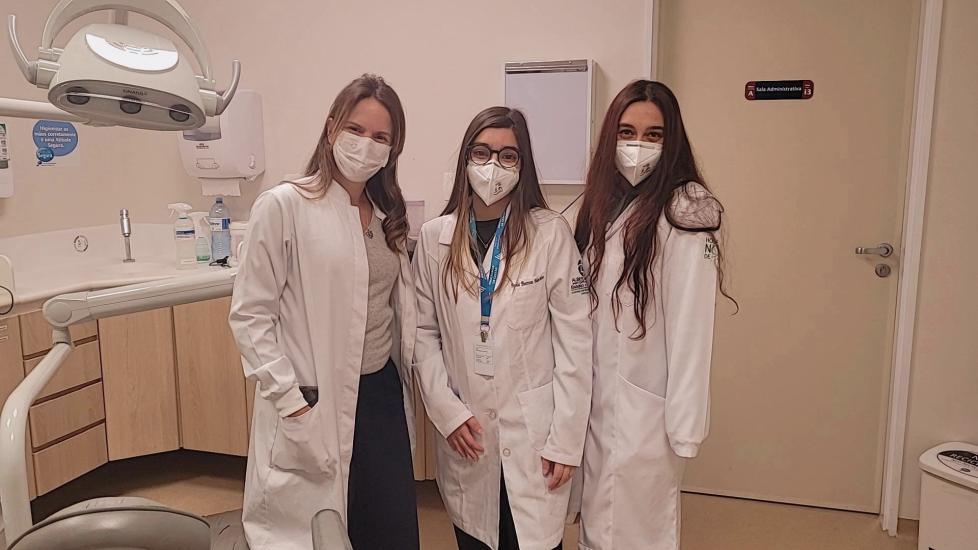 Prathysha Kothare standing far right with two of her colleagues in white lab coats in a Hospital Israelita Albert Einstein room with a white corner desk and some sanitization sprays on top of it behind them