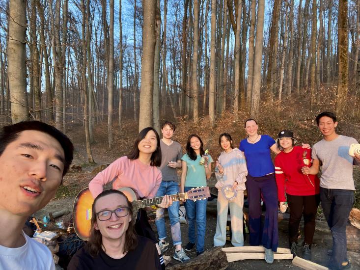 Raphi Kang with a guitar in hand pictured in a selfie with 8 other friends in a forest surrounded by trees during sunset