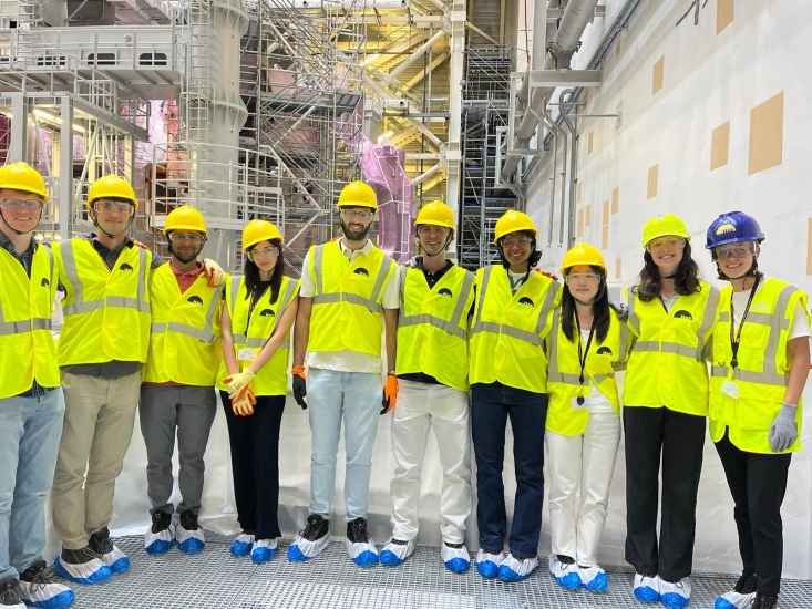 Dilin Melon and nine colleagues in neon yellow vests and caps with protective shoe gear in a lab in France. Behind him and his colleagues are scaffoldings