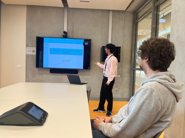 Ishan Ganguly dressed formally in a dress shirt presenting his work in the front of the room while his colleague in grey sweatshirt listens in