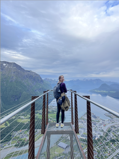Ashley Margetts standing on a mountain viewing platform with a scenic view of mountains, water, and a town below.