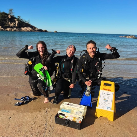 Andrew Motz holding one of his flipper while squatting on the beach in a divers suit with two others