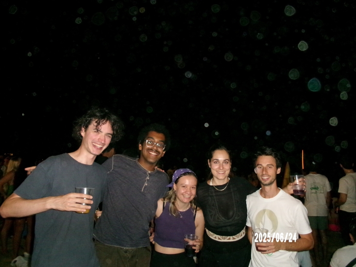 Caleb (second from left) with four other colleagues holding drinks and arms around each other shoulders at night