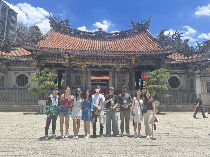 Achival Goel, pictured third from right, and 8 others in front of a traditional temple with a red-tiled roof and intricate carvings.