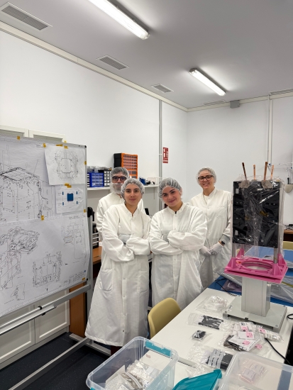 Emily standing with crossed arms wearing a full lab coat with hair cap next to a white board with 3 lab mates