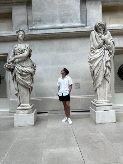 Kenny with both hands in his shorts' pockets looking up at two towering marbled sculptures draped in togas
