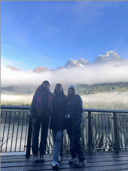 Mina Cezairli (left) and two friends standing in front of a fence with a snowcapped mountainous region behind clouds