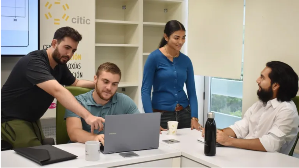 Jace Lopez seated in front of a laptop while a colleague standing next to him pointing at the laptop. Two other colleagues talking on next to Jace
