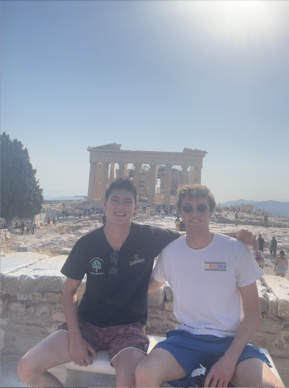 Sean Huckleberry seated on a stone bench with an arm around a friend to his right on a sunny day with the Parthenon in Athens, Greece in the background