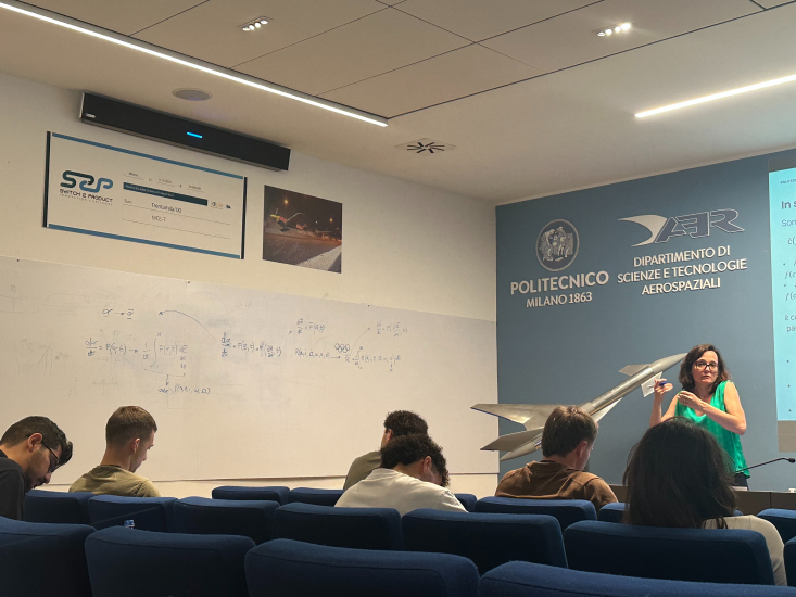 A person in green giving a talk in a room with a whiteboard to the left and a screen to the right with blue cushioned chairs