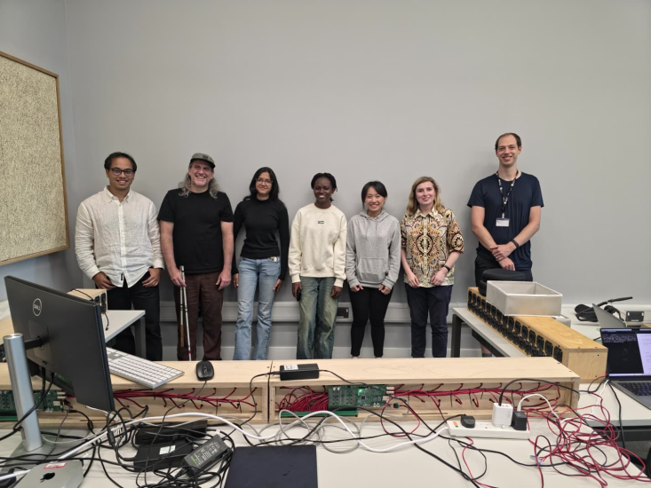 Riya S with six colleagues standing against a white wall with a table in the front with red wiring